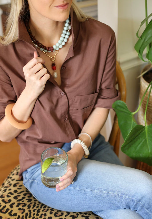 Woman sitting with a glass of water, wearing a brown shirt and blue jeans, with a plant in the background.