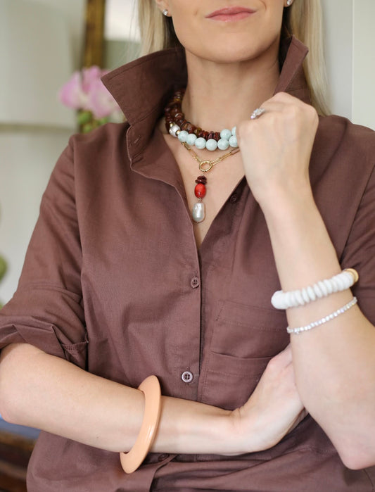 Woman wearing a brown shirt with layered necklaces and bracelets indoors.