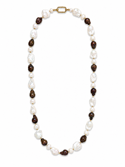 A woman wearing a long beaded necklace with irregular white, cream, and gray beads, paired with a brown sleeveless top, set against a softly blurred outdoor background.